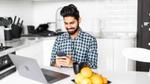 A young man with his phone and laptop in a white kitchen with bright yellow lemons on top