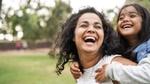 A woman with curly hair smiles while carrying a child on their back, set against a green park background.