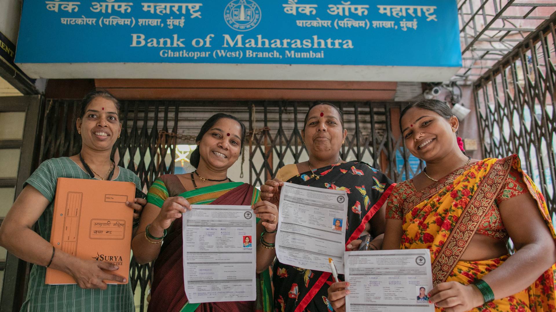 Four smiling Indian women showcase their Bank of Maharashtra loan documents outside the Ghatkopar branch in Mumbai, highlighting financial inclusion and women's empowerment.