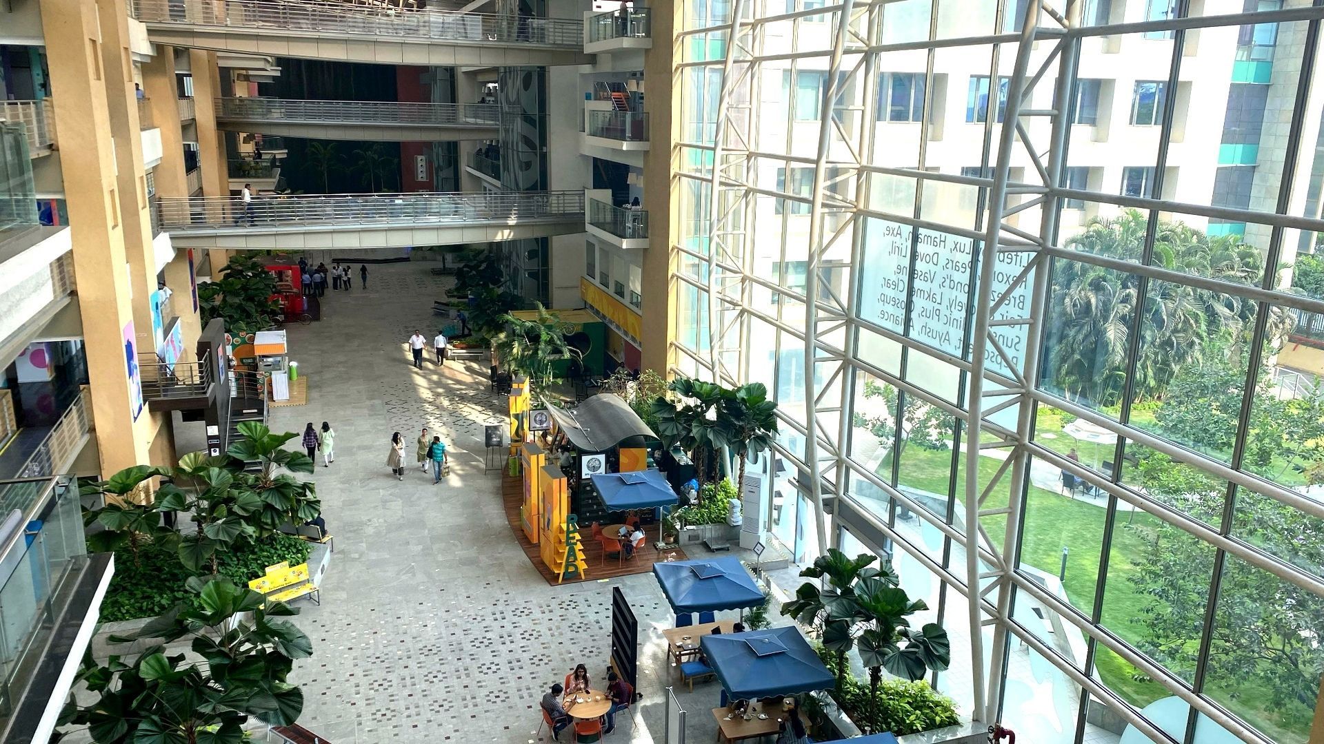 People walk and dine in a spacious, plant-filled corporate atrium with floor-to-ceiling windows.