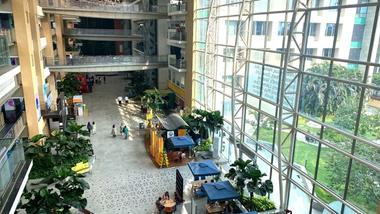 People walk and dine in a spacious, plant-filled corporate atrium with floor-to-ceiling windows.
