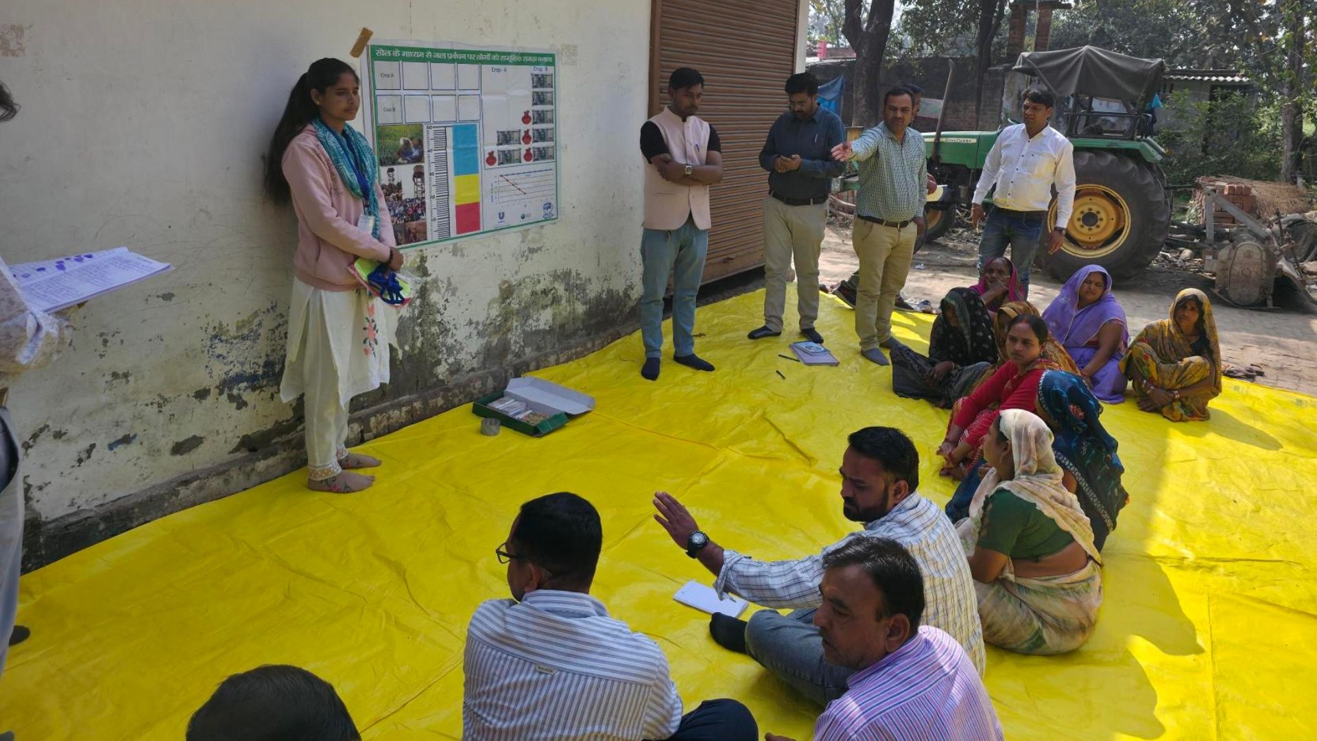 Facilitator leads an outdoor training session with villagers seated on a yellow mat