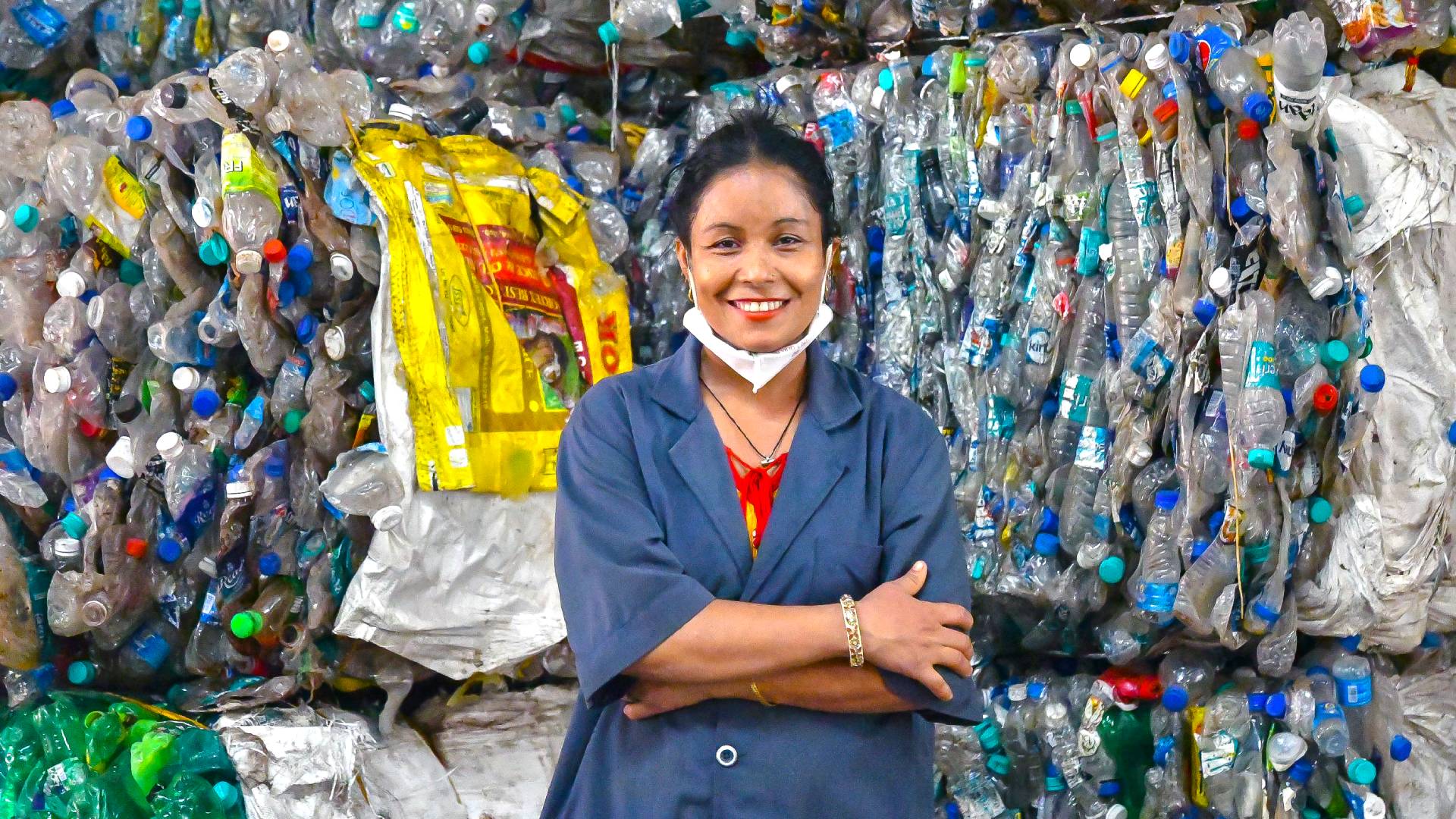 Smiling worker stands in front of a wall of compacted, recycled plastic bottles and packaging materials, promoting sustainable recycling practices.