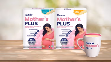 Two packs of Horlicks Mother's Plus nutrition drink in vanilla and kesar flavours featuring a pregnant woman in a pink dress, along with a filled mug displayed on a wooden surface.