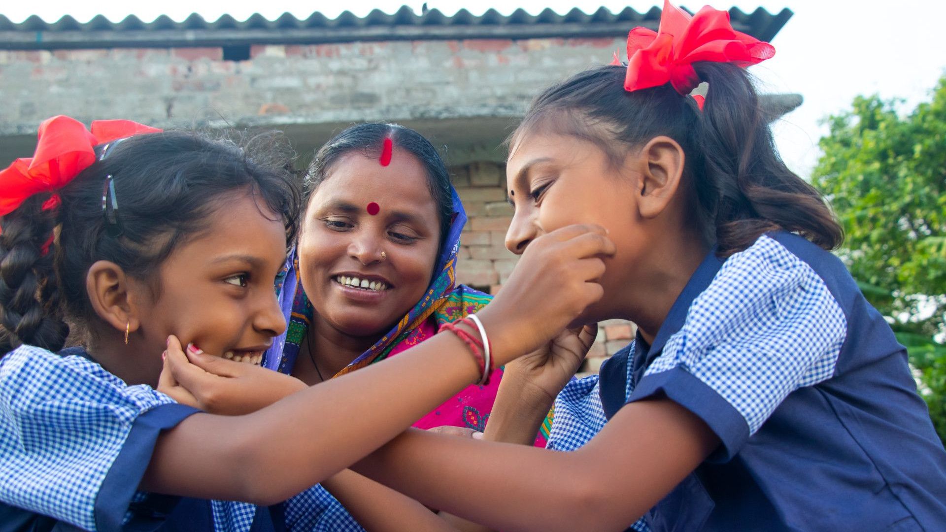 Three girls in school uniforms with red bows engage playfully outdoors, surrounded by greenery and a brick wall backdrop.