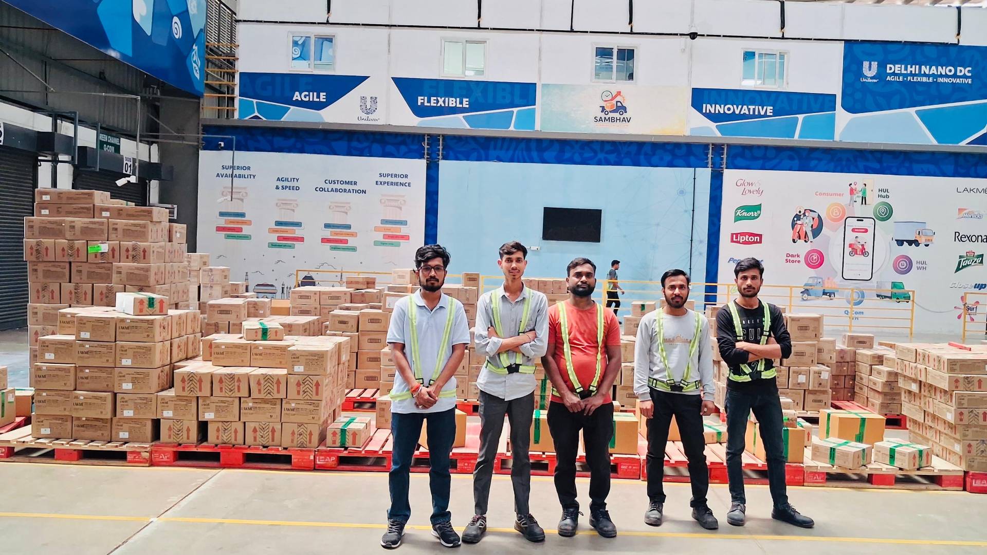 Five warehouse workers posing in front of stacked boxes.