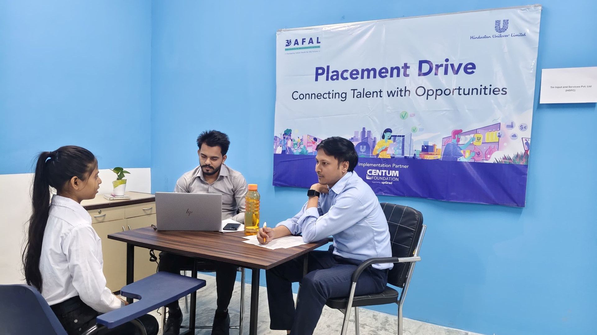Three people in a room with a laptop and documents on the table, under a banner reading "Placement Drive".