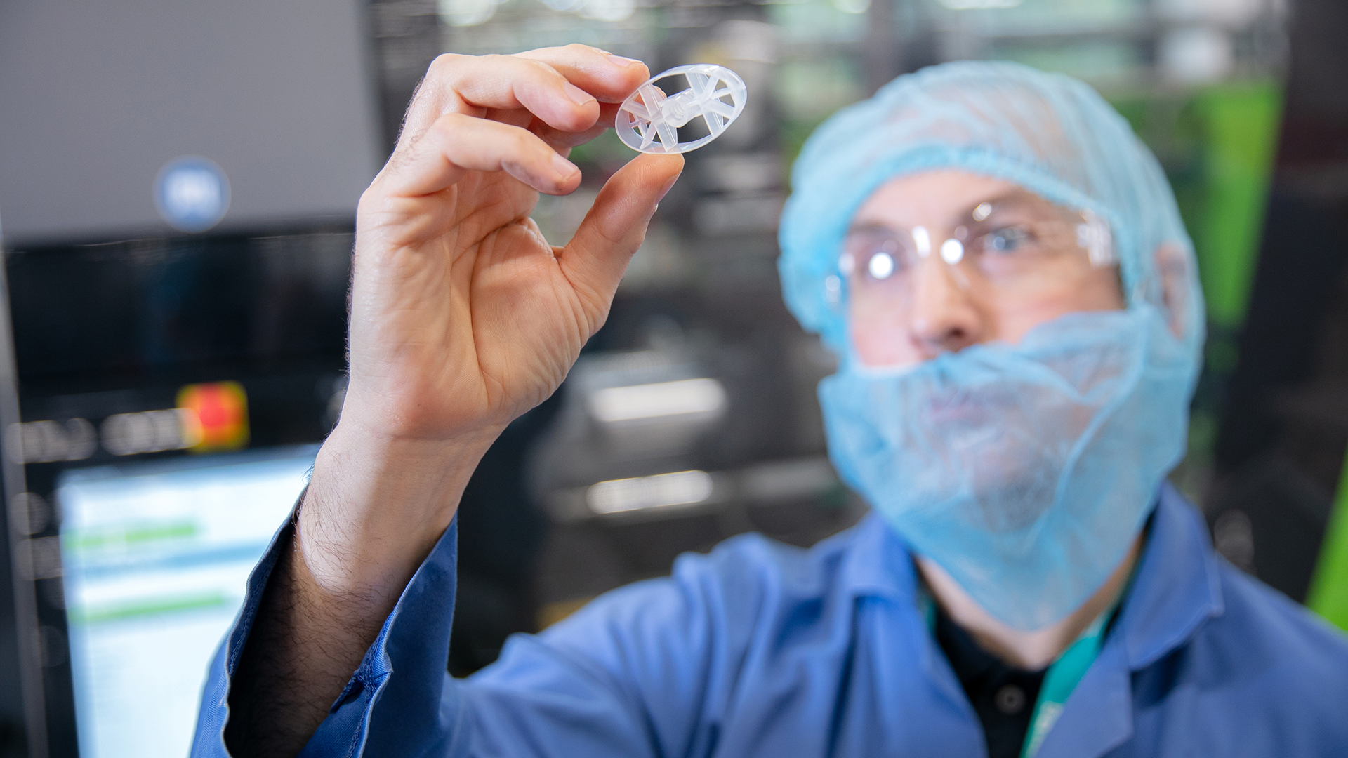 A gloved hand holding a small, circular plastic component in a cleanroom environment with machinery in the background.