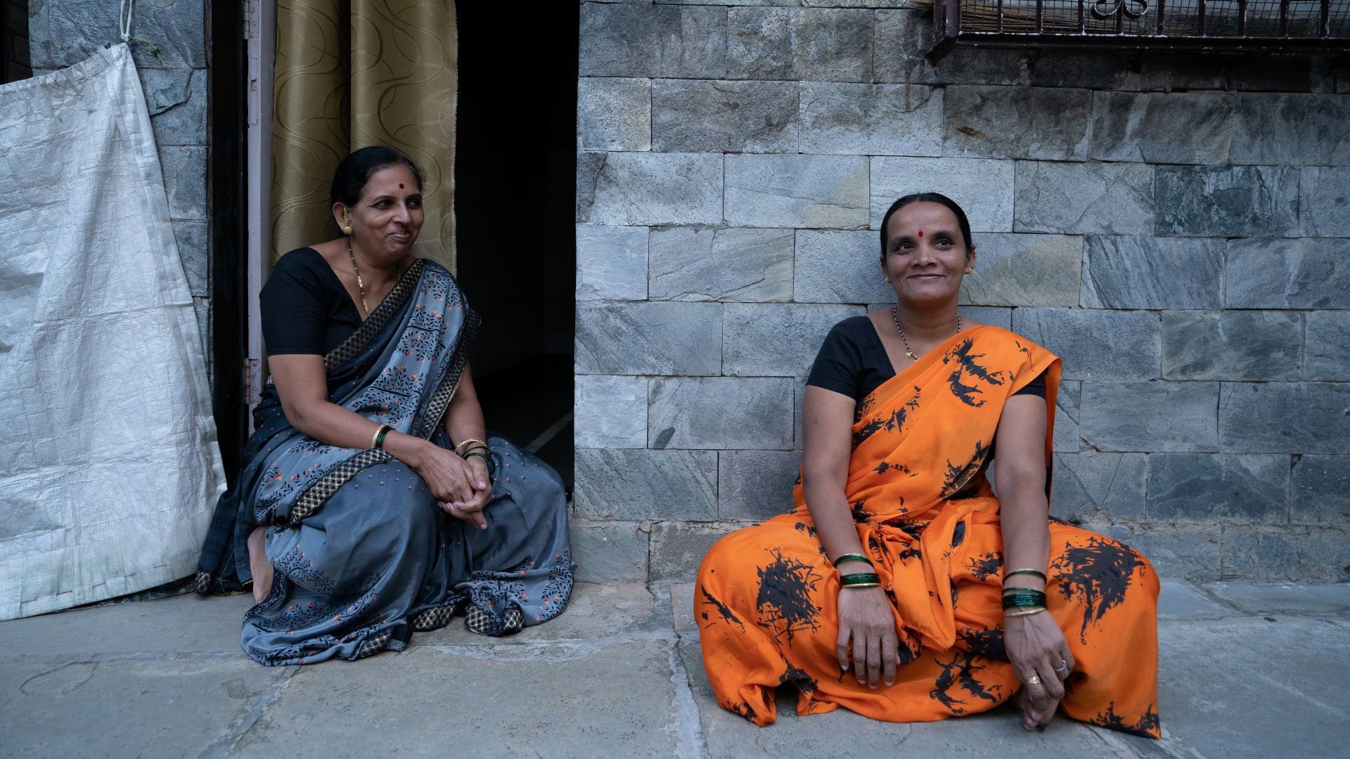 Two women in colorful saris sit outside a stone-walled building.