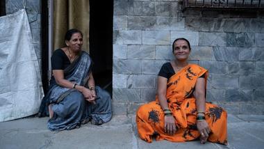 Two women in colorful saris sit outside a stone-walled building.
