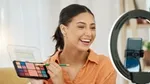 A woman in orange shirt holding a makeup palette and a brush filming herself with a phone and a ring light.