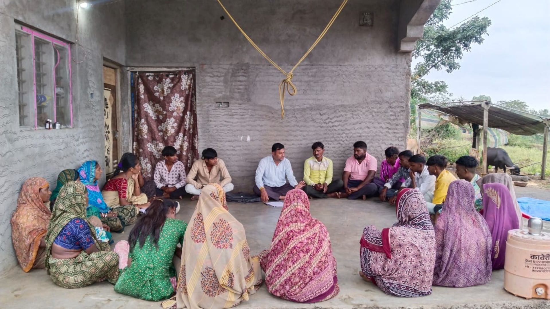 Community members sit in a circle for a group discussion outside a rural home.
