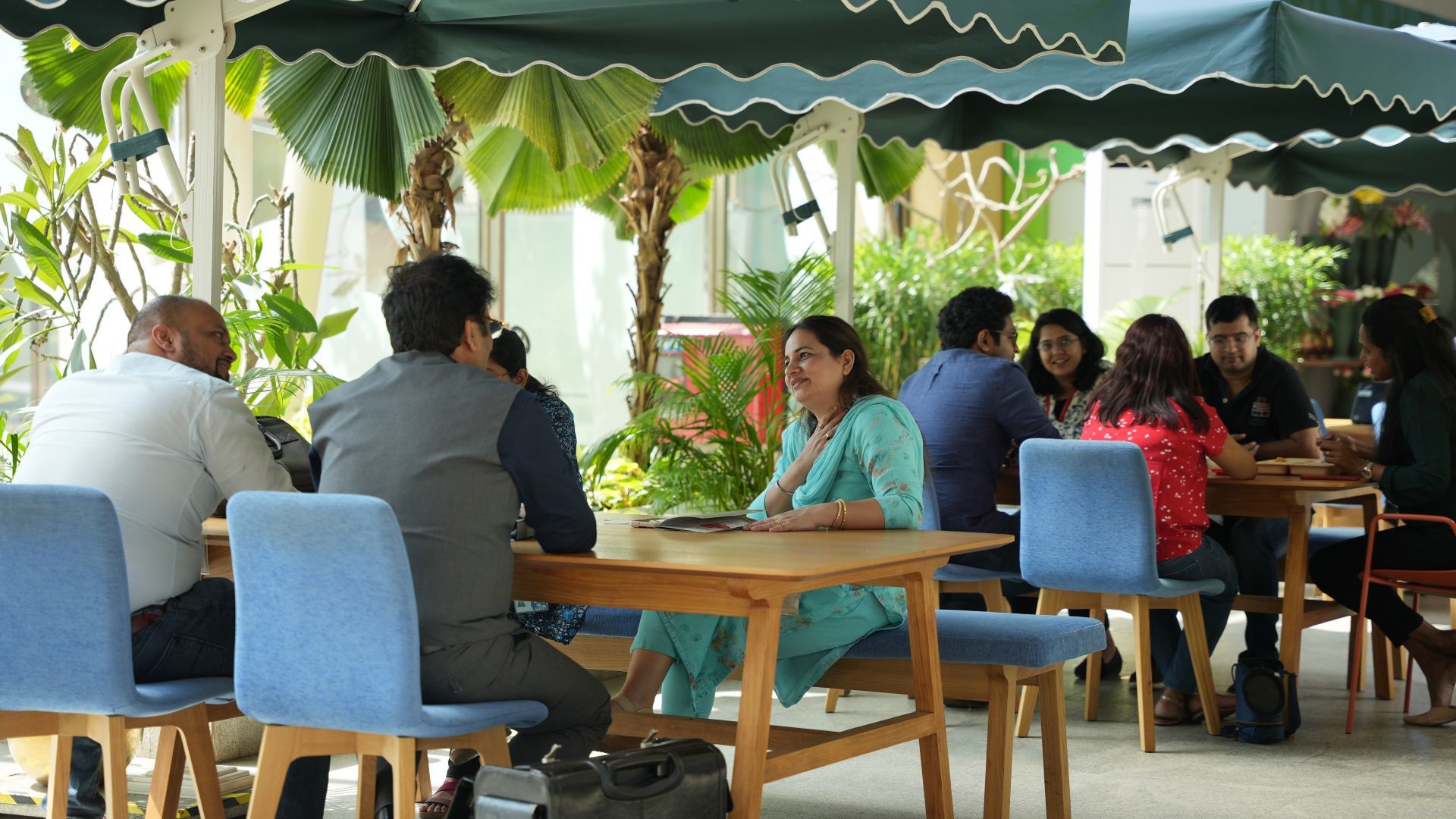 People sit and converse at wooden tables under green and white canopies in a lush outdoor cafe setting.