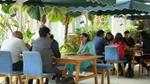 People sit and converse at wooden tables under green and white canopies in a lush outdoor cafe setting.