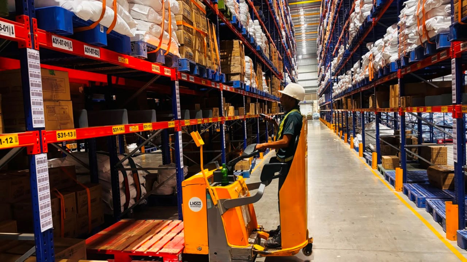 Warehouse worker on a pallet truck navigating tall racks filled with organized inventory.