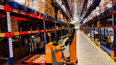 Warehouse worker on a pallet truck navigating tall racks filled with organized inventory.