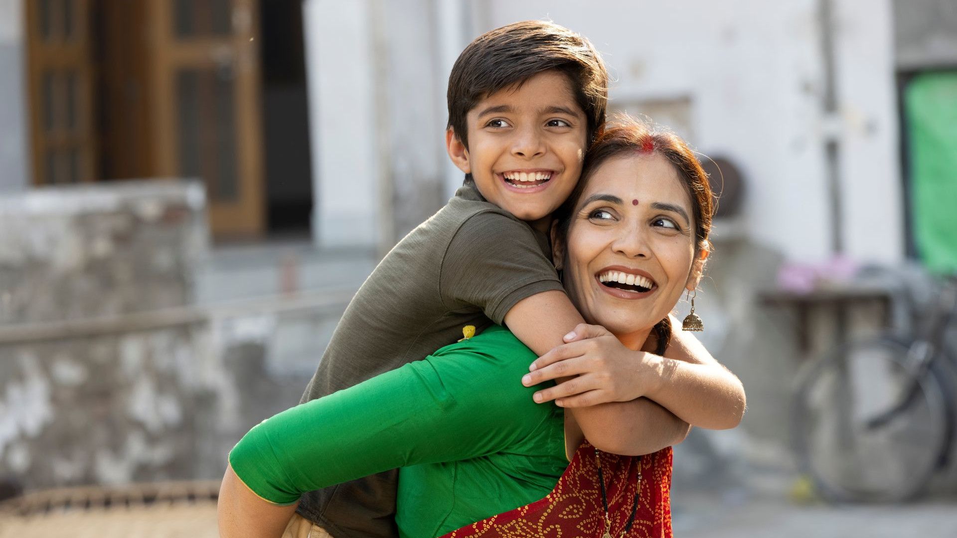 A child playfully hugs an adult wearing vibrant clothing in a sunny outdoor setting.