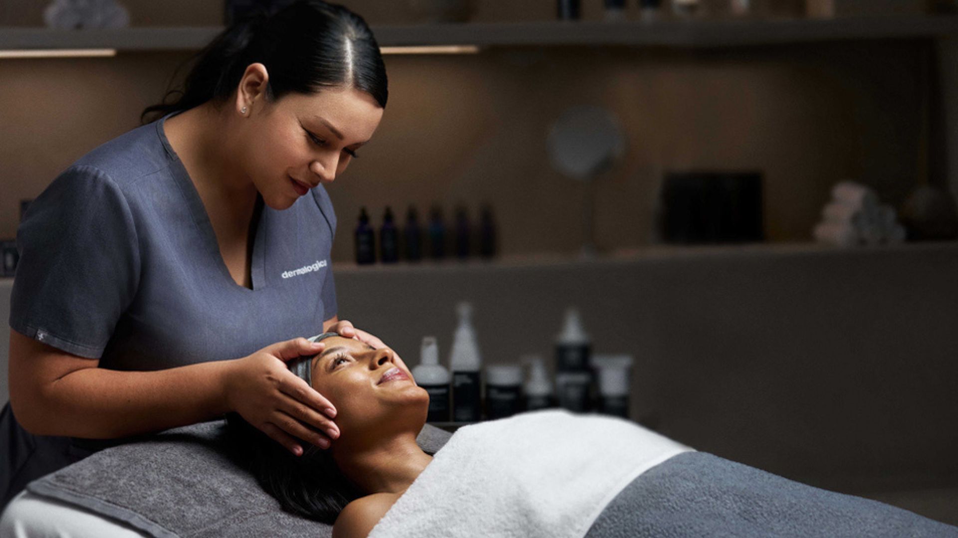 Spa therapist gently massages a client’s face in a treatment room with skincare products in the background