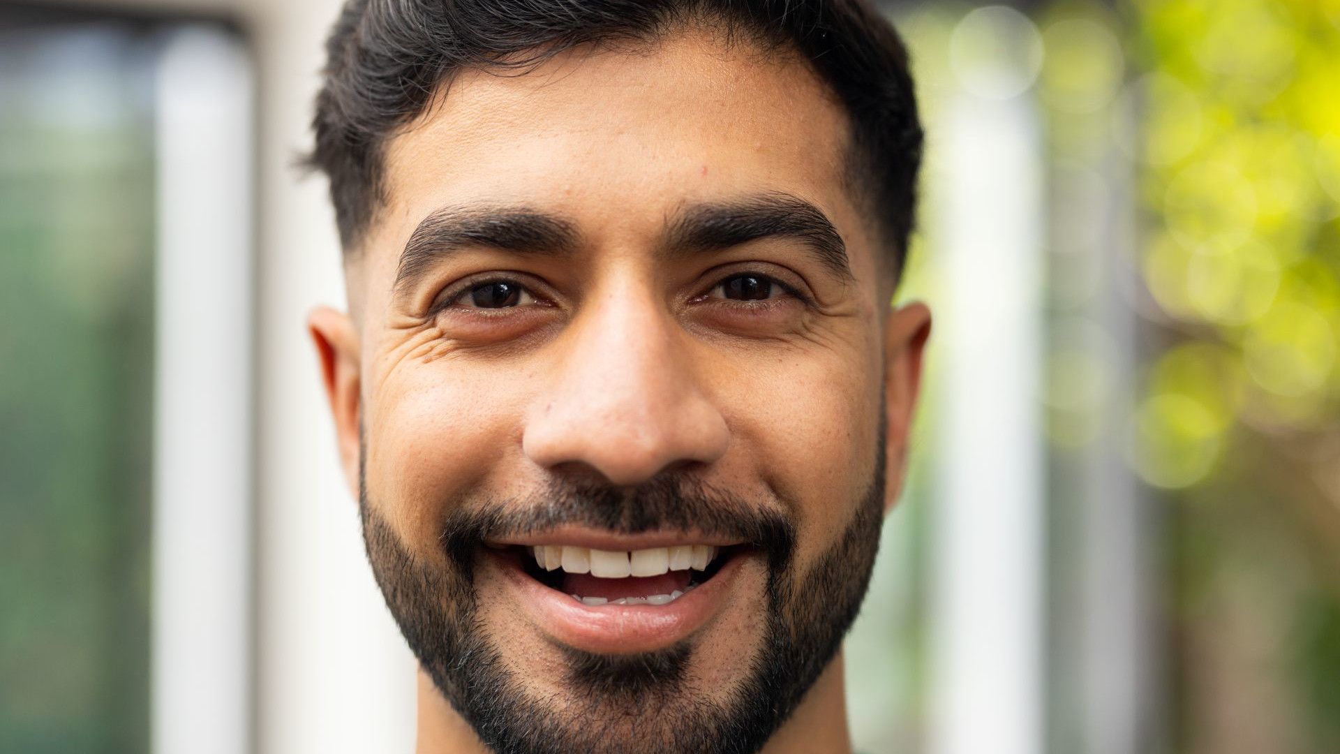 A close-up portrait of a young man with dark hair, standing in front of a blurred outdoor background filled with greenery.