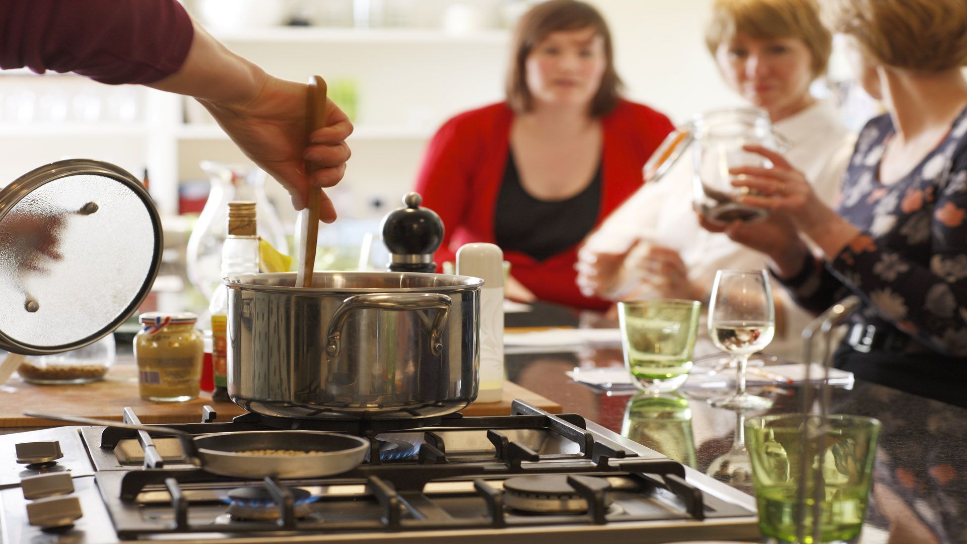 A kitchen scene, with a person stirring a pot on a hob and three women looking at ingredients