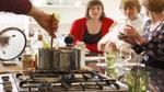 A kitchen scene, with a person stirring a pot on a hob and three women looking at ingredients
