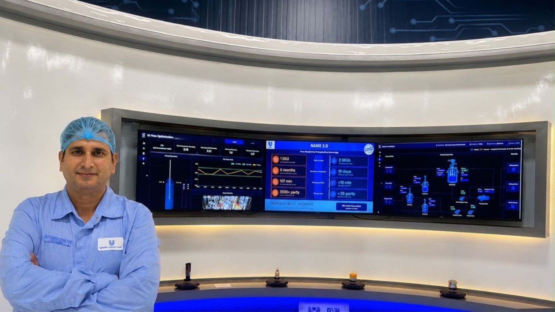 Factory engineer in protective gear stands before large digital dashboards showing production analytics.