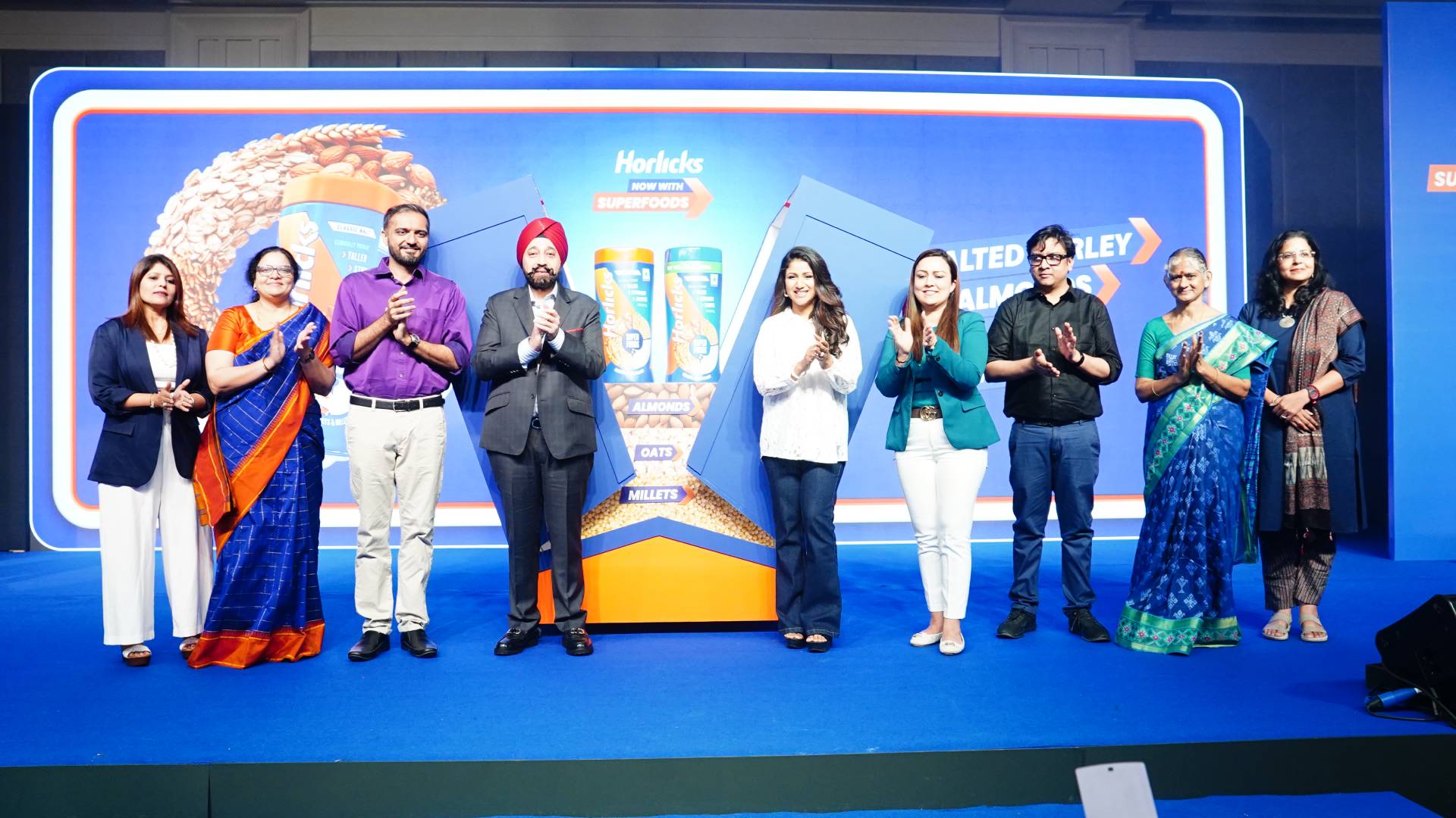 Group of people clapping on stage with Horlicks banner in the background.