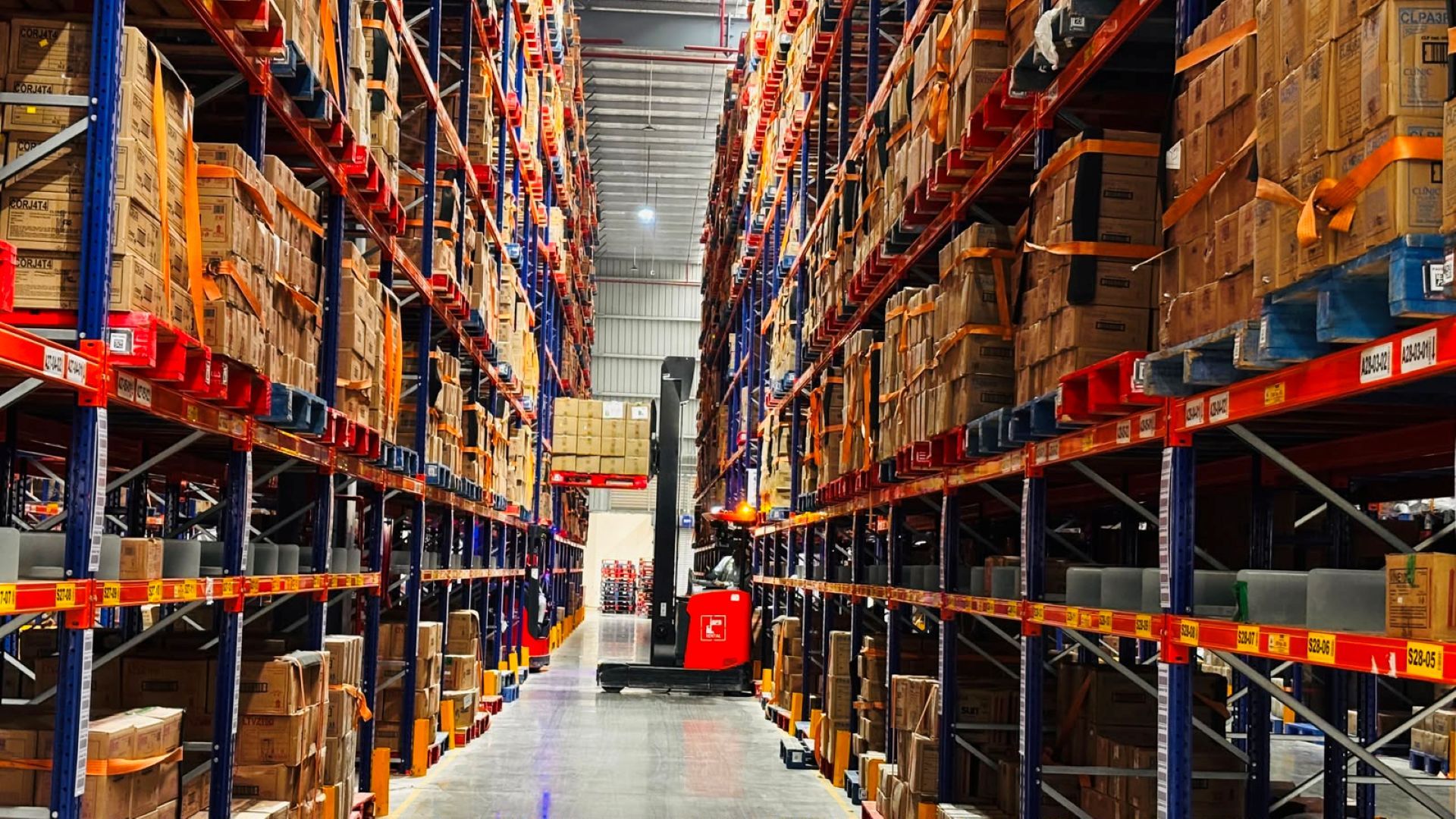 Forklift moves pallets between tall racks of boxed inventory in a brightly lit warehouse aisle.