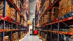 Forklift moves pallets between tall racks of boxed inventory in a brightly lit warehouse aisle.