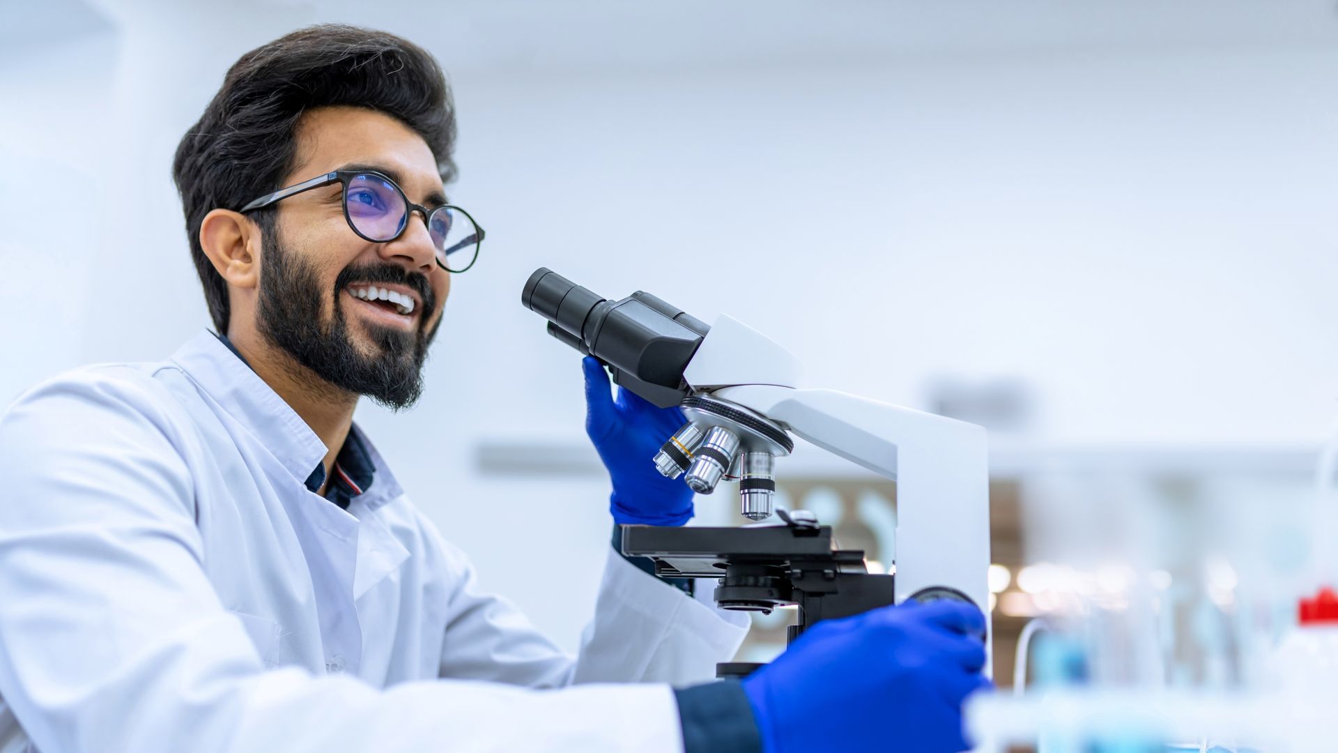 A man in a lab coat and blue gloves examines a sample under a microscope, surrounded by laboratory equipment.