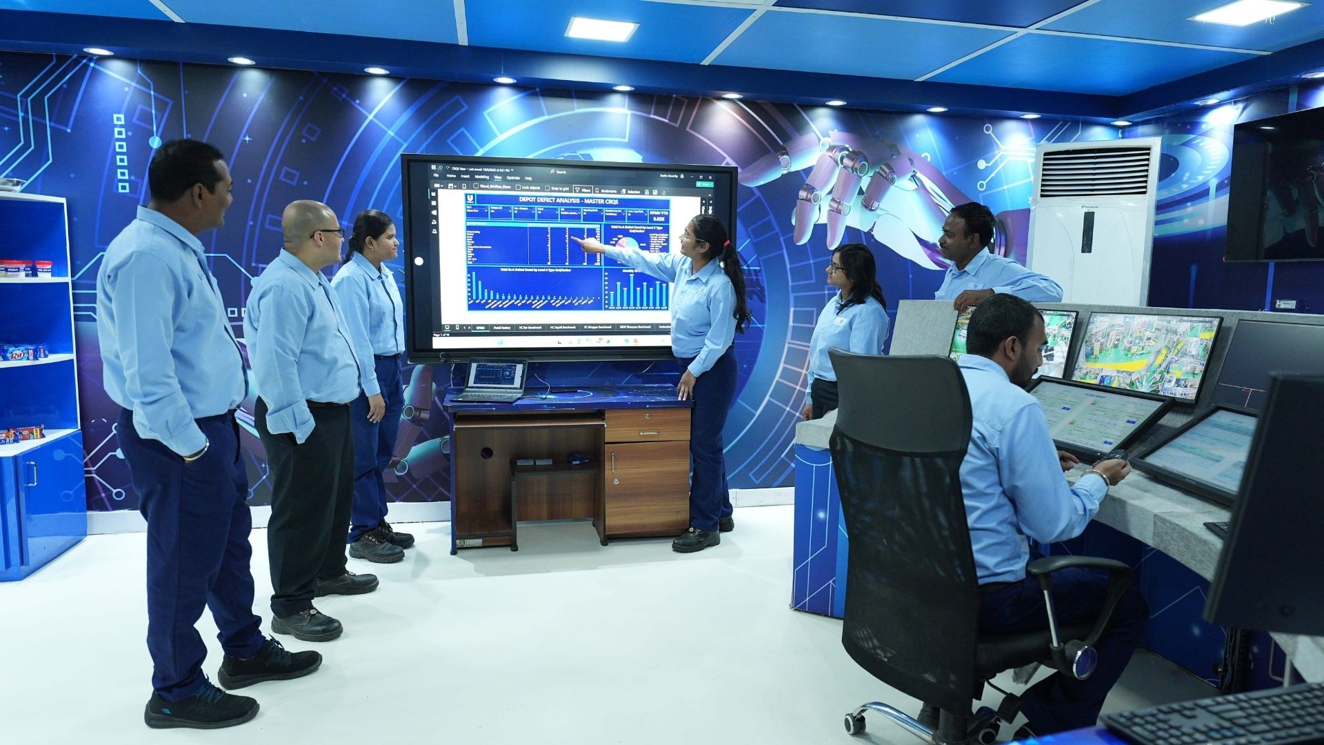 People in a control room with monitors, discussing data on a large screen.