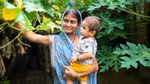 A Prabhat beneficiary alongside her nutri garden. The beneficiary is a lady in blue saree and is holding a baby in her hand.