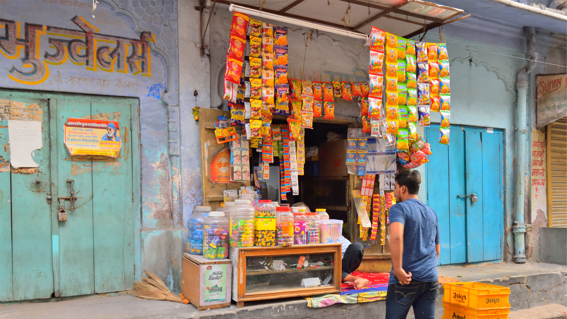 A small street-side shop displaying hanging packets of snacks and jars of candies, with a person standing in front of the shop.