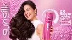 A woman with thick, flowing brown hair smiles, holding a pink Sunsilk shampoo bottle.