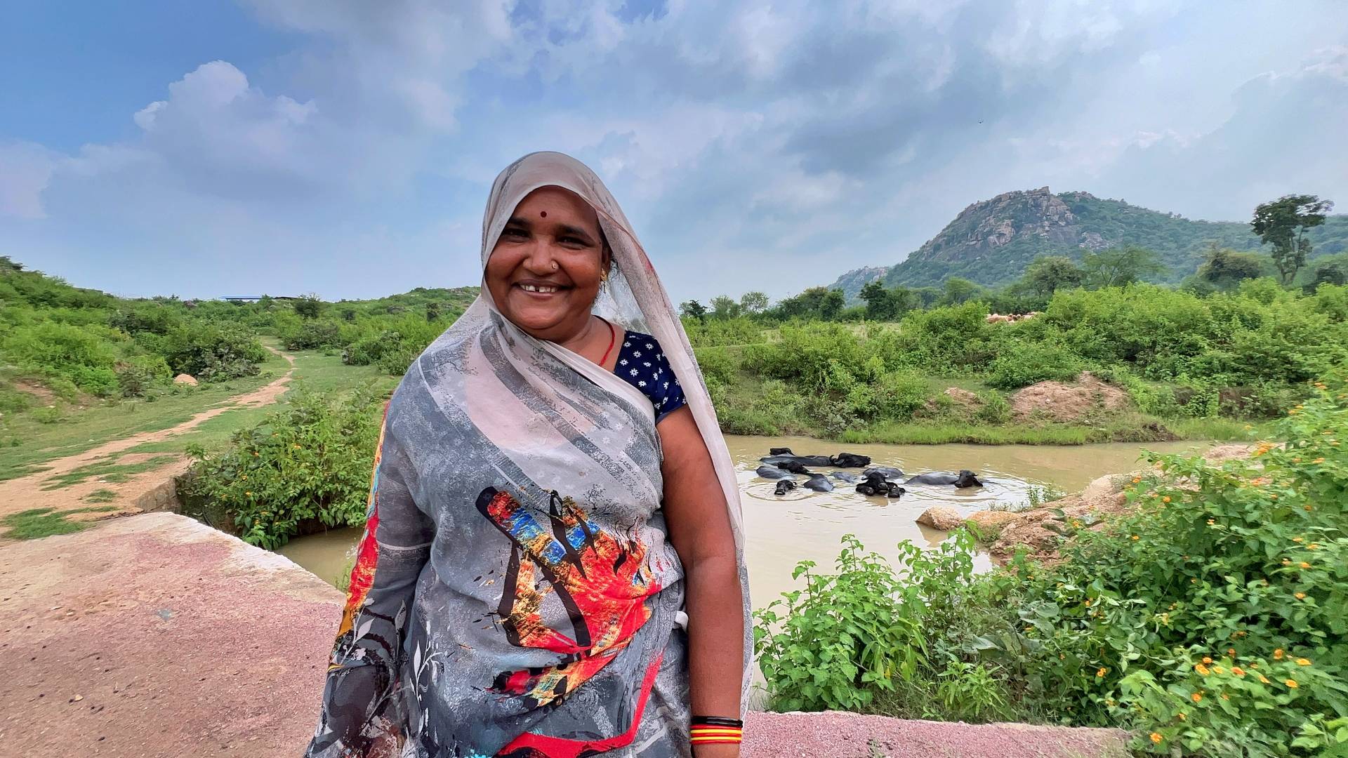Smiling woman in colorful saree near a pond with lush greenery and a hill in the background.