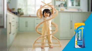 A child in a kitchen is surrounded by a DNA-shaped swirl of grains, symbolizing growth, with a Junior Horlicks container on the right.