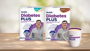 Two packs of Horlicks Diabetes Plus in vanilla and chocolate flavours featuring a smiling older man, along with a Diabetes Plus branded mug displayed on a wooden surface.