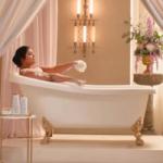 A woman relaxing in a vintage clawfoot bathtub surrounded by soft pink curtains, elegant lighting, and a floral arrangement.