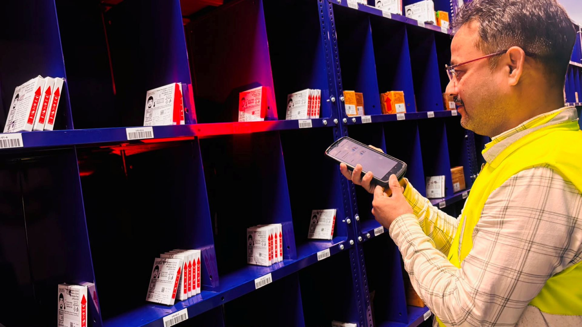 Warehouse worker in a safety vest scans labeled boxes on blue shelves with a handheld device.