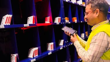 Warehouse worker in a safety vest scans labeled boxes on blue shelves with a handheld device.