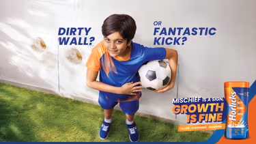 A child in a blue and orange outfit holds a soccer ball, standing next to a wall with stains and graphic text