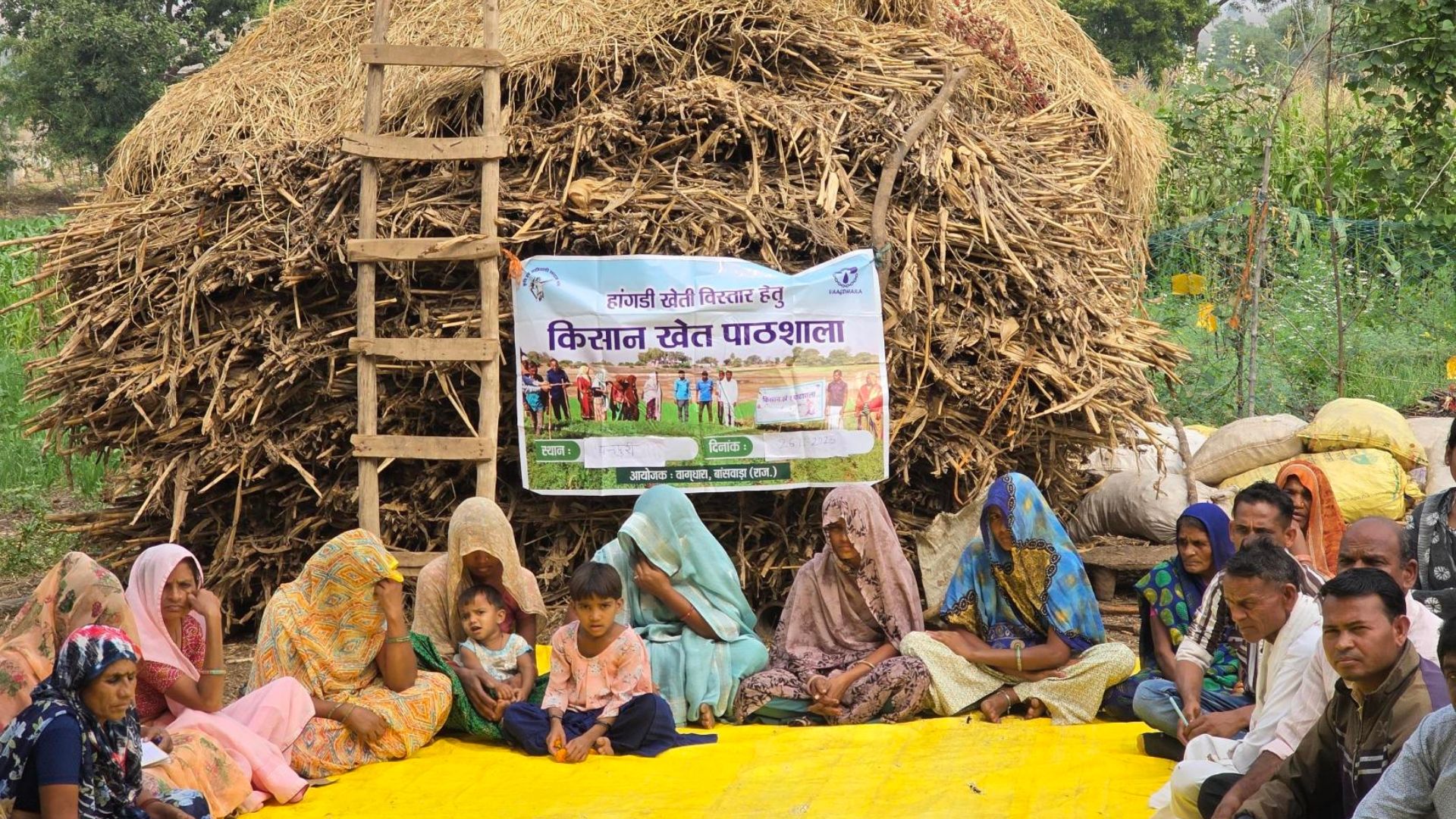 Rural community members sit on a mat near a haystack for an outdoor farmer training session.