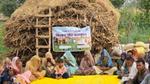 Rural community members sit on a mat near a haystack for an outdoor farmer training session.
