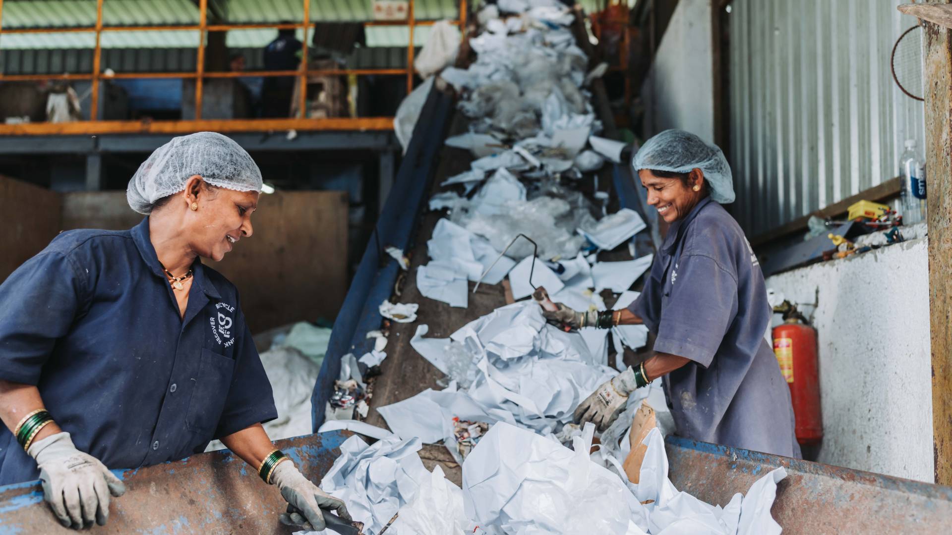 Two women workers sorting recyclable plastic waste on a conveyor belt at a recycling plant.