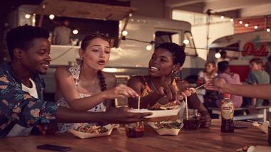 Three people sitting at a table in front of takeaway food cartons, drinking Lipton Ice Tea.