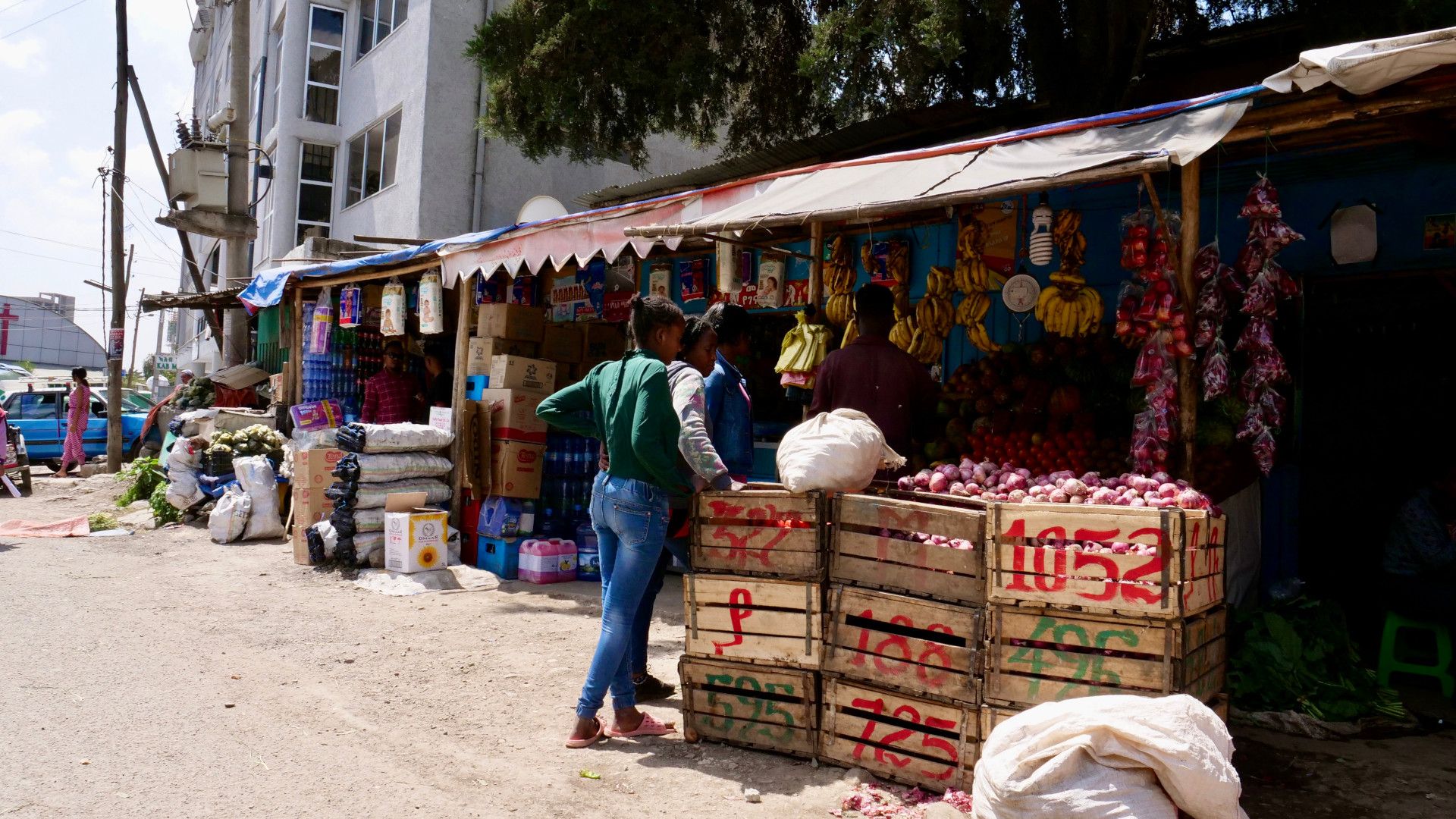 A woman shops at a market in rural India. Fruit is hanging overhead. Boxes full of vegetables.