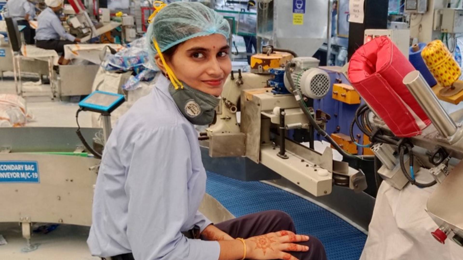 Worker in uniform and hairnet seated near machinery in factory.