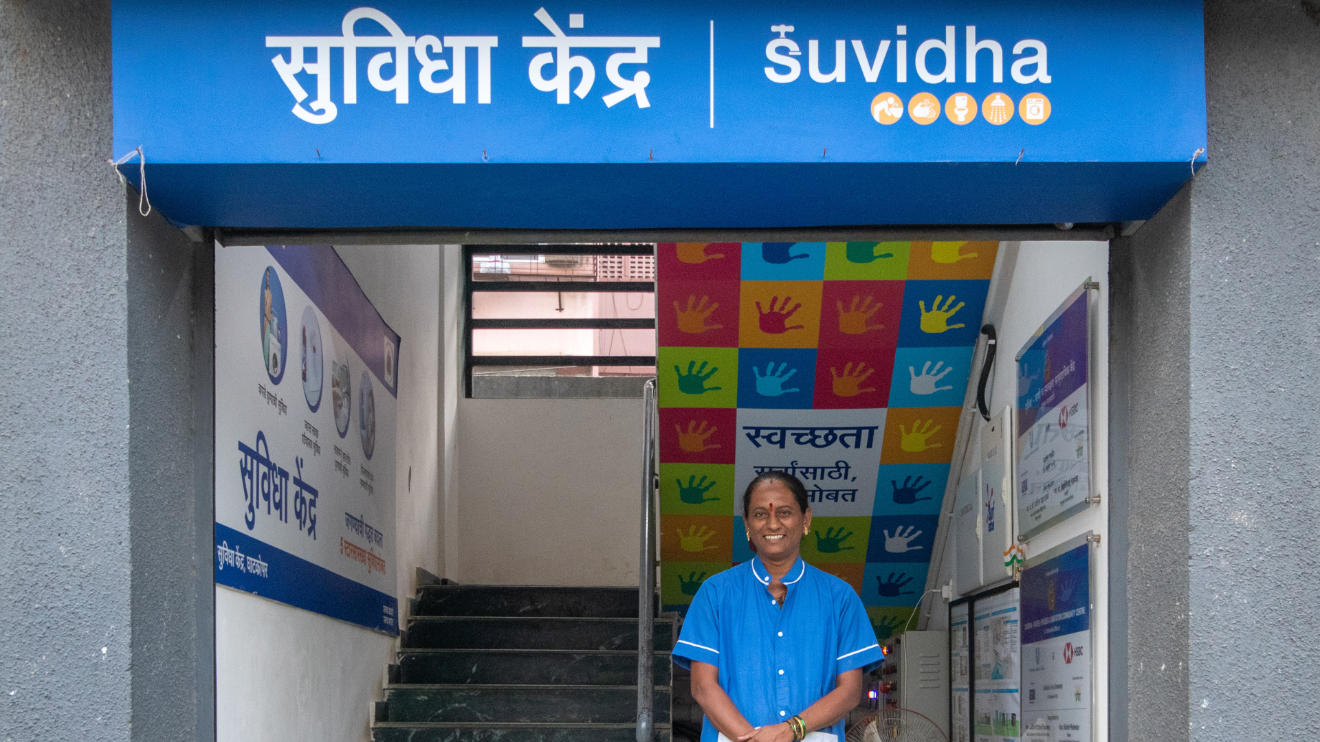 A woman in blue shirt standing and smiling with the Suvidha signboard above her head