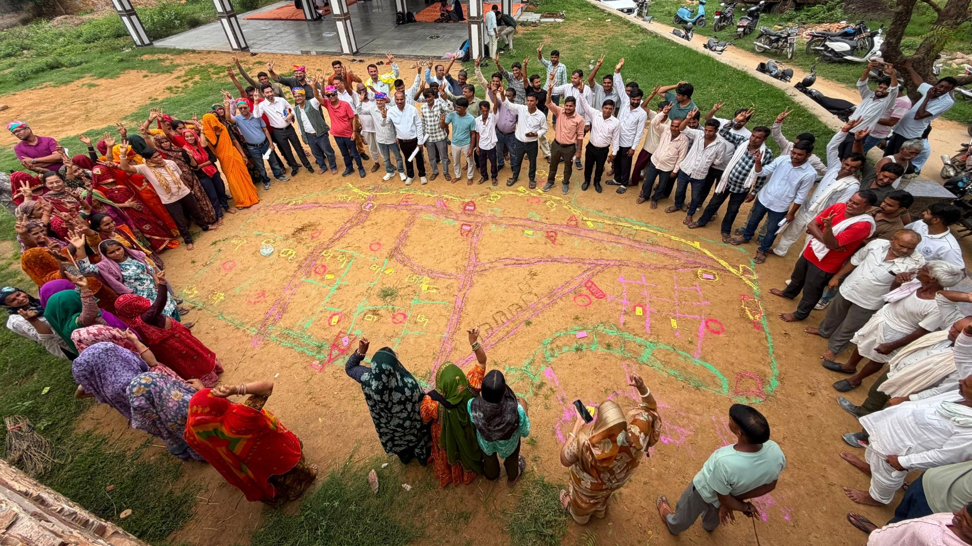 Community members stand in a circle outdoors, discussing a colorful map drawn on the ground.