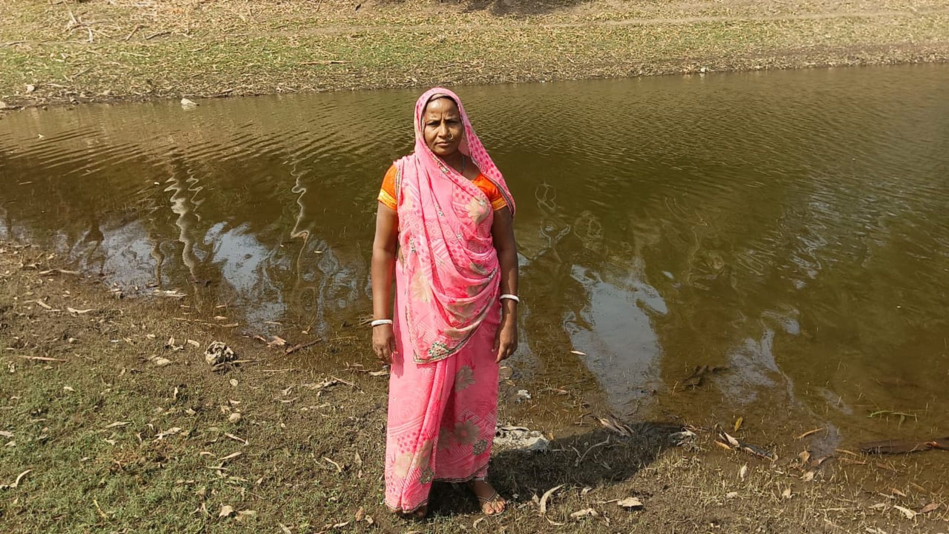 Woman in a pink sari stands beside a rural pond.